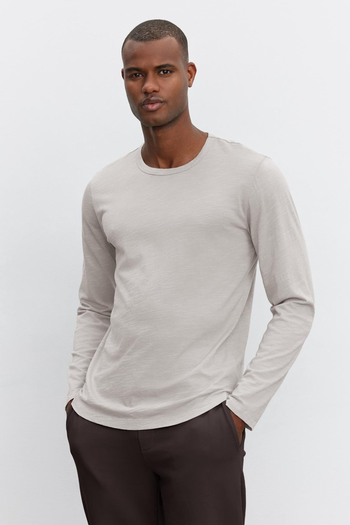 A person wearing the KAI TEE by Velvet by Graham & Spencer, a light gray long-sleeve shirt crafted from lightweight Peruvian cotton with a classic crew neckline, stands in front of a plain white background while paired with dark pants. The shirt features a timeless design.-38264832131265