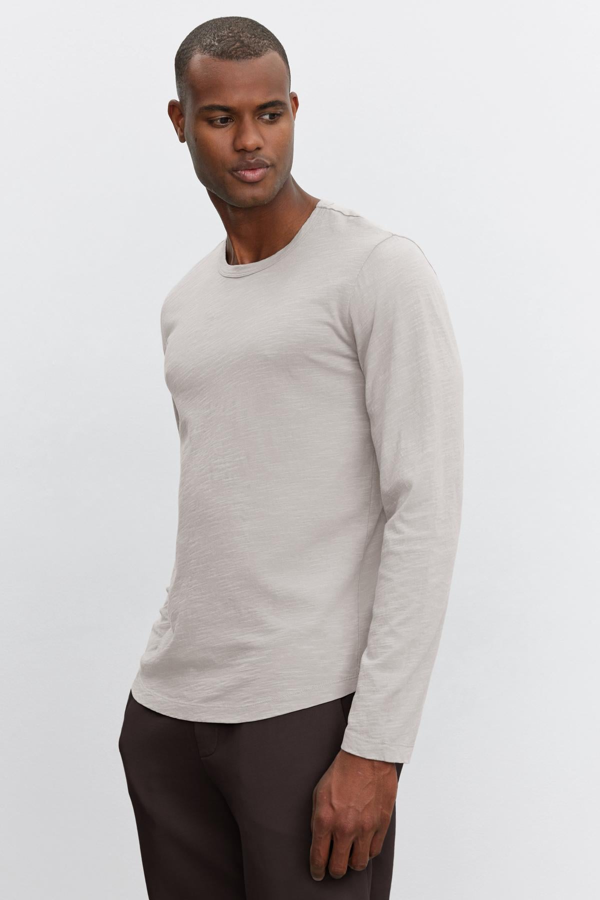   A person wearing the sophisticated KAI TEE from Velvet by Graham & Spencer, showcasing a light gray long-sleeve shirt crafted from luxurious Peruvian cotton paired with dark pants, stands against a plain white background. 