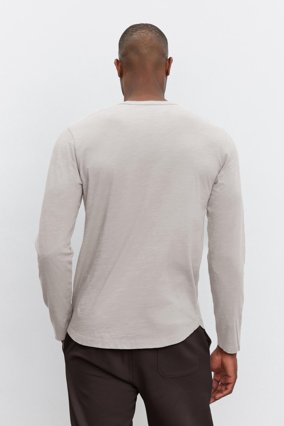   The individual is depicted from behind against a plain background, wearing the KAI TEE, a classic light gray long-sleeve shirt made from lightweight Peruvian cotton by Velvet by Graham & Spencer, paired with dark pants. 