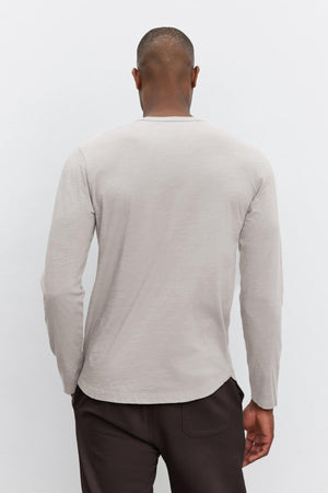 The individual is depicted from behind against a plain background, wearing the KAI TEE, a classic light gray long-sleeve shirt made from lightweight Peruvian cotton by Velvet by Graham & Spencer, paired with dark pants.