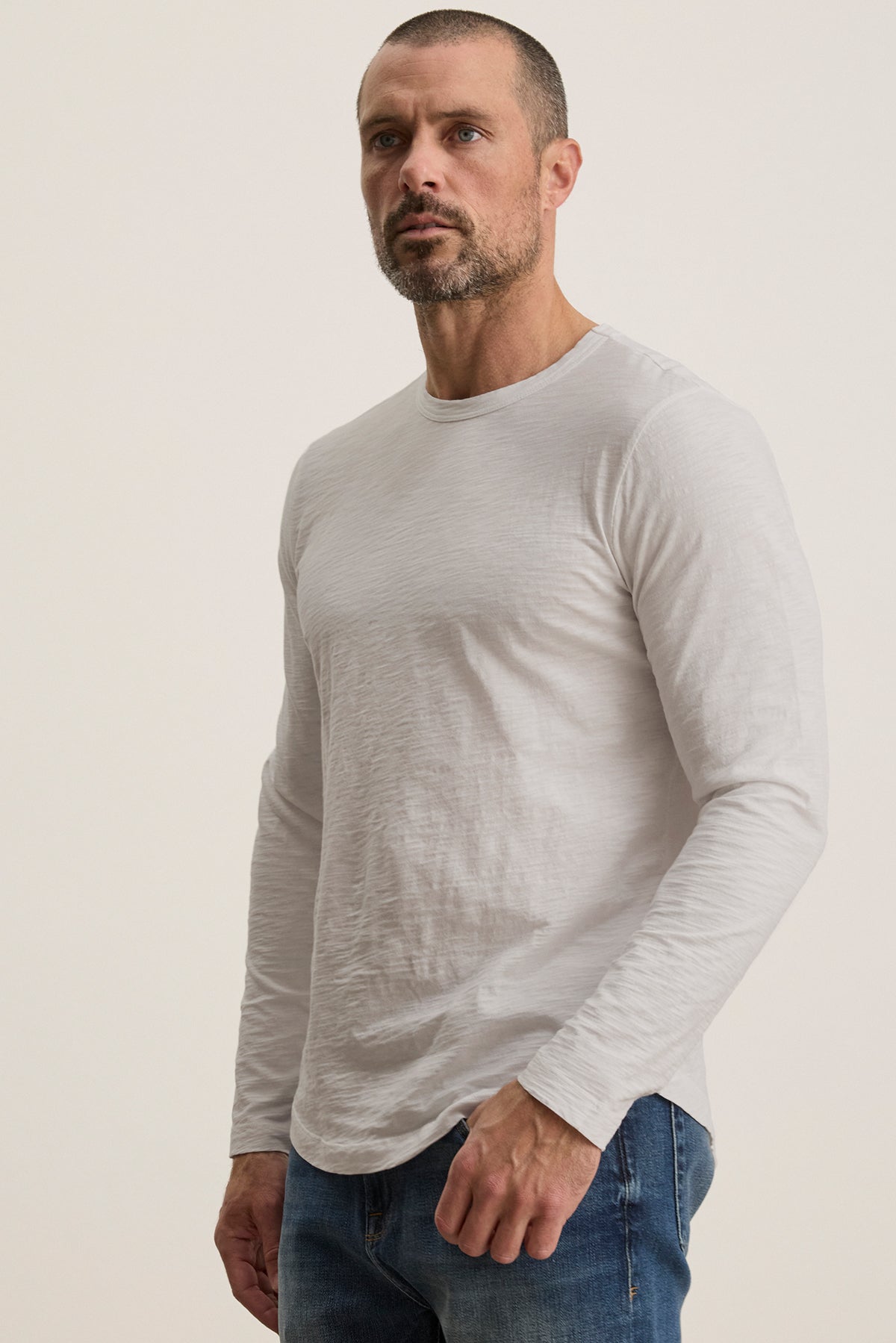   A bearded man with short hair wears the KAI TEE by Velvet by Graham & Spencer, a plain white long-sleeve shirt with a crew neckline and timeless design, paired with jeans against a neutral background. 