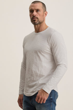 A bearded man with short hair wears the KAI TEE by Velvet by Graham & Spencer, a plain white long-sleeve shirt with a crew neckline and timeless design, paired with jeans against a neutral background.