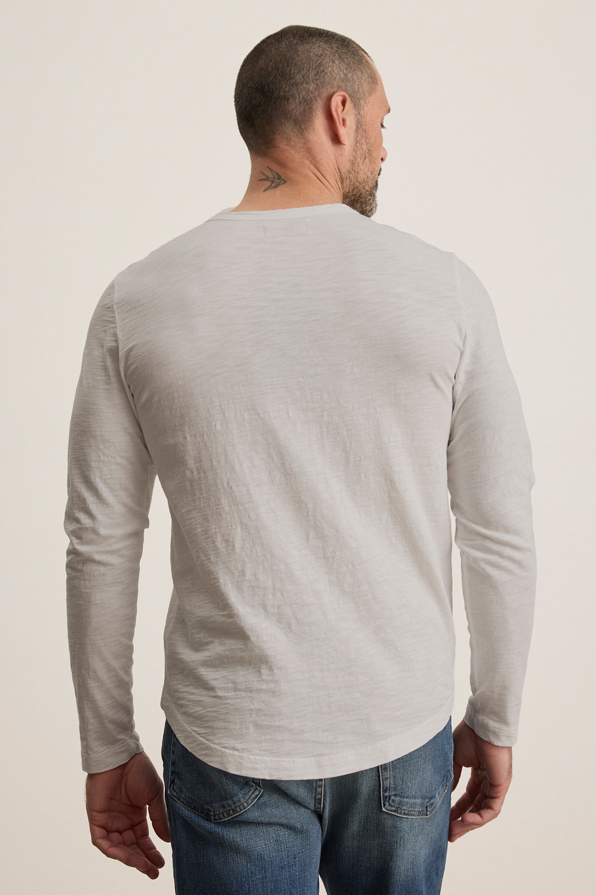   A man with short hair and a small neck tattoo, dressed in a KAI TEE by Velvet by Graham & Spencer—a white long-sleeve shirt with a crew neckline and timeless design—paired with jeans, is facing away from the camera. 