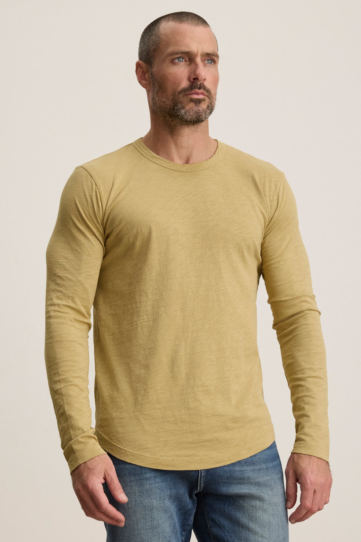 A man with short hair and a beard wears the KAI TEE by Velvet by Graham & Spencer—a light yellow Peruvian cotton long sleeve crewneck tee—paired with blue jeans, standing against a plain light background.-39957108621505