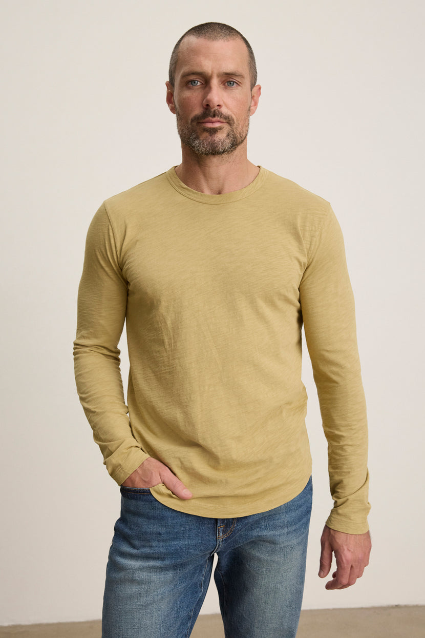 A man wearing the KAI TEE by Velvet by Graham & Spencer—a mustard yellow long sleeve crewneck—and blue jeans stands against a plain, light background with one hand in his pocket.