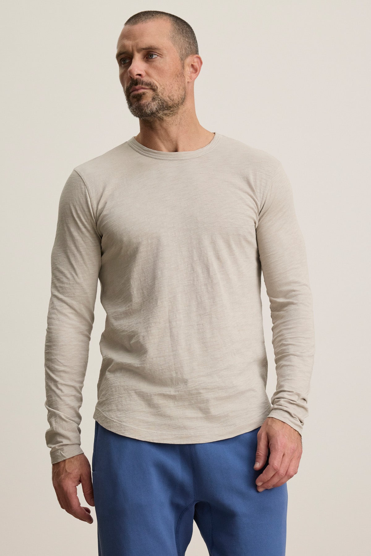 A man stands slightly to the side against a plain background, wearing the KAI TEE by Velvet by Graham & Spencer—a beige long-sleeve crew neck tee—paired with blue pants.