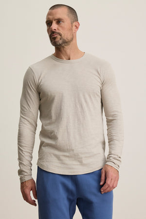 A man stands slightly to the side against a plain background, wearing the KAI TEE by Velvet by Graham & Spencer—a beige long-sleeve crew neck tee—paired with blue pants.