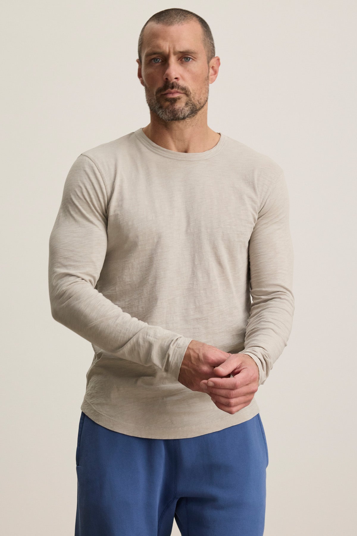 A man with short hair and a beard wears the KAI TEE by Velvet by Graham & Spencer—a light beige crewneck Peruvian cotton tee—with blue pants, standing against a plain, light background.-40452483186881