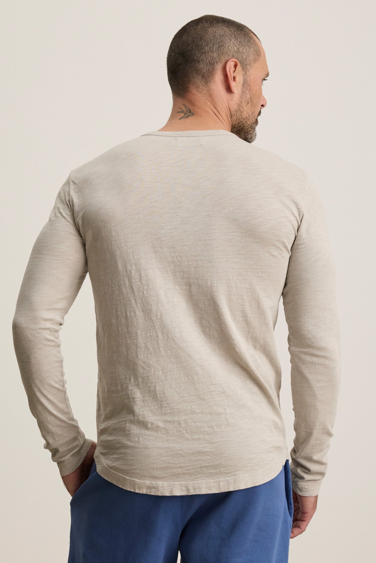 A man with a short haircut and neck tattoo is seen from the back, wearing a beige Velvet by Graham & Spencer KAI TEE and blue pants against a plain background.-40452483121345