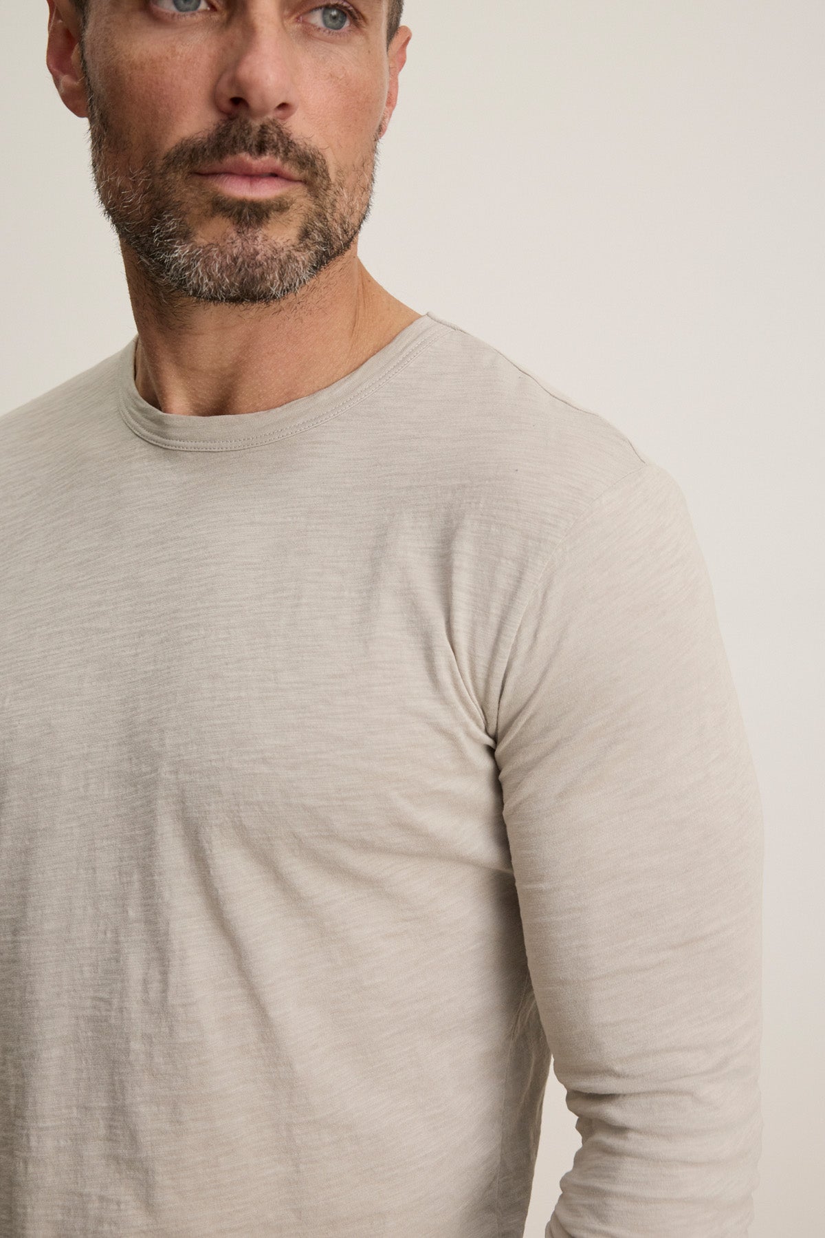 A bearded man wears the Velvet by Graham & Spencer KAI TEE—a long sleeve, crew neck tee in light beige Peruvian cotton—shown from the chest up against a plain background.
