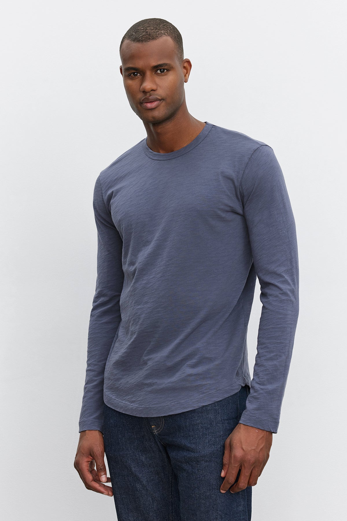   A man stands against a plain background, wearing a long-sleeve, dark grey KAI TEE from Velvet by Graham & Spencer and jeans. 