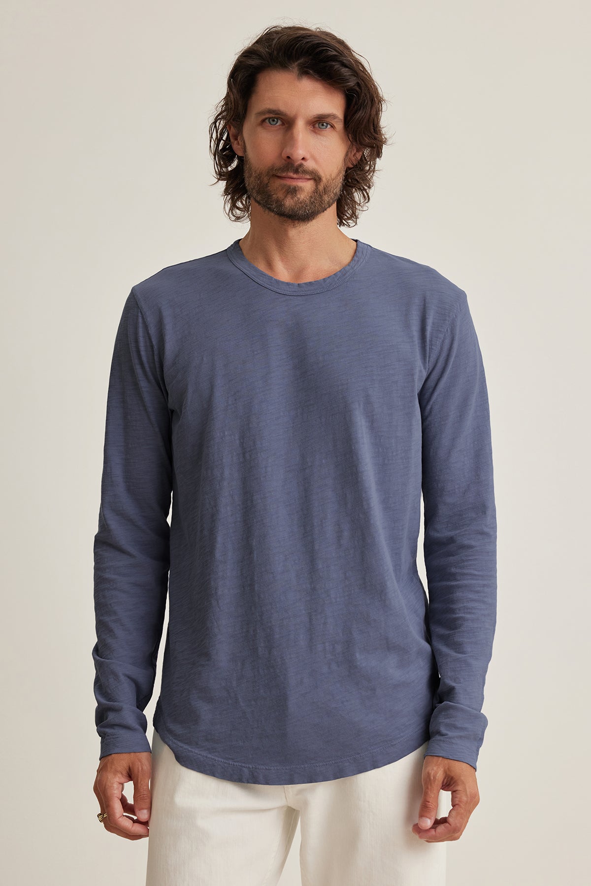   Wearing a KAI TEE by Velvet by Graham & Spencer, made of soft Peruvian cotton, a person with shoulder-length hair stands against a neutral background. The blue long-sleeve shirt, paired with white pants, features a classic crew neckline for a timeless look. 