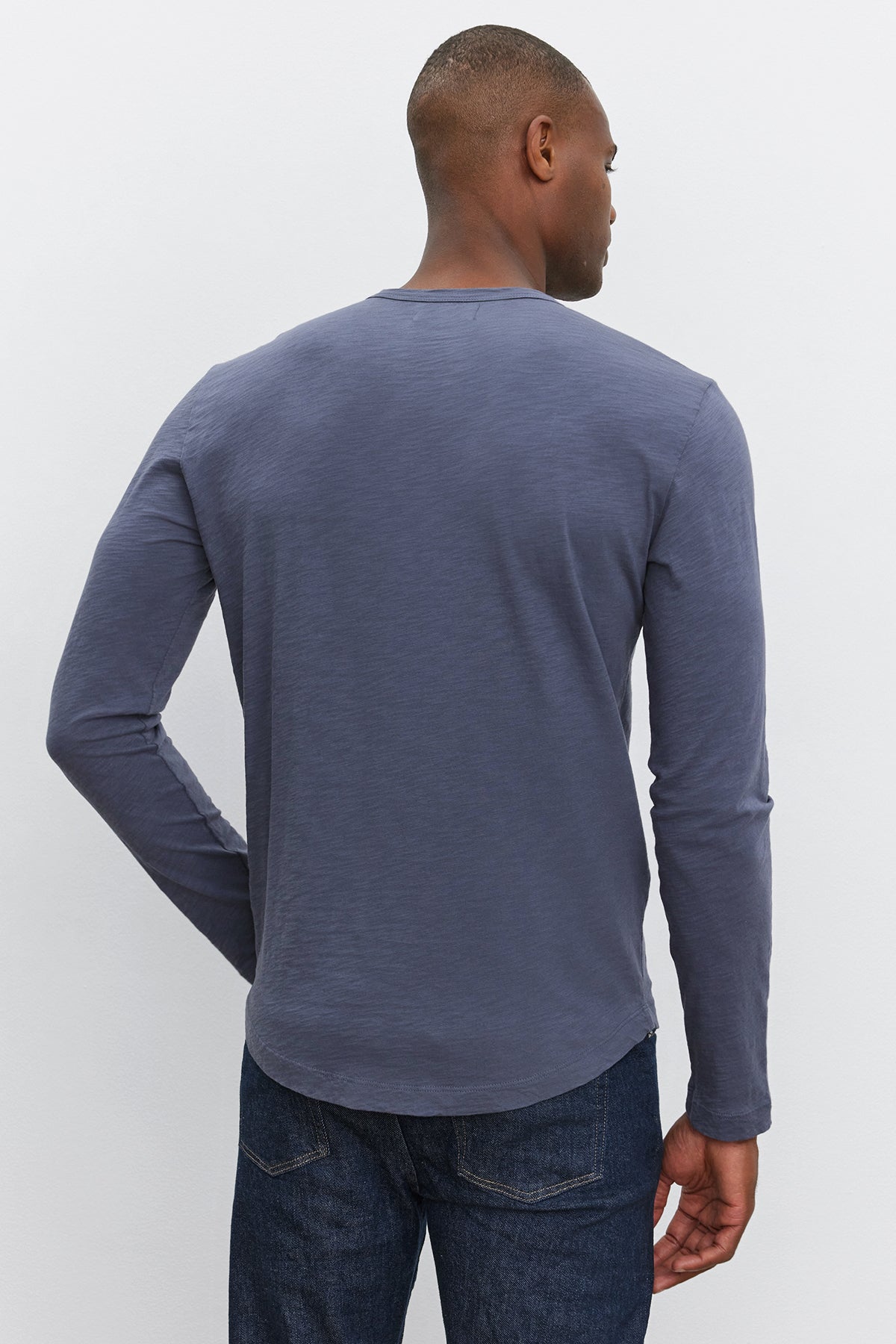 A man is standing with his back to the camera, wearing the Velvet by Graham & Spencer KAI TEE, a long-sleeved dark gray shirt with a classic crew neckline, paired with dark blue jeans against a light-colored background.-37893903253697