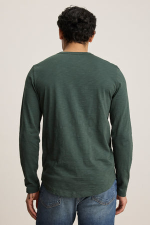 A man is shown from the back against a plain background wearing the Velvet by Graham & Spencer KAI TEE, a long-sleeve dark green Peruvian cotton crewneck, paired with blue jeans.