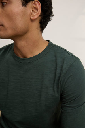 Wearing the KAI TEE by Velvet by Graham & Spencer—a dark green Peruvian cotton tee with a classic crew neckline—this person is seated, facing slightly left, with only their upper torso and part of their face visible.
