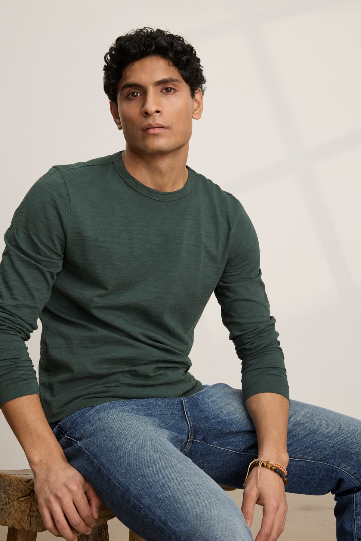 A man with short curly hair, wearing the Velvet by Graham & Spencer KAI TEE—a green Peruvian cotton long sleeve crew neck shirt—and blue jeans, sits on a wooden stool against a plain light background.-39905774796993