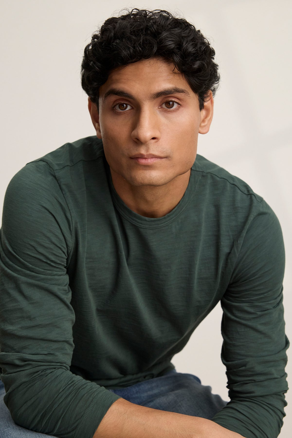 A man with short curly dark hair wears the Velvet by Graham & Spencer KAI TEE, a dark green Peruvian cotton long sleeve crewneck, and looks at the camera with a neutral expression against a plain background.-39905774698689