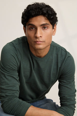 A man with short curly dark hair wears the Velvet by Graham & Spencer KAI TEE, a dark green Peruvian cotton long sleeve crewneck, and looks at the camera with a neutral expression against a plain background.
