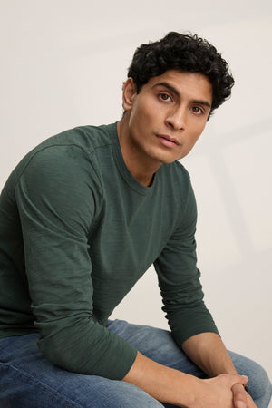 A man with short curly hair wears the KAI TEE by Velvet by Graham & Spencer—a green Peruvian cotton crewneck—paired with jeans as he sits and gazes past the camera against a light background.