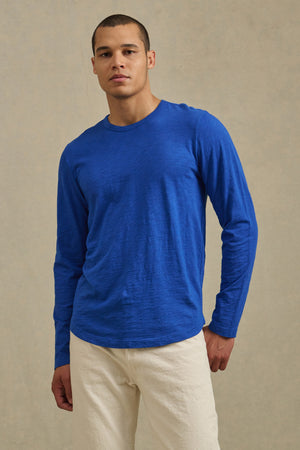 A man stands against a plain background wearing the Velvet by Graham & Spencer KAI TEE in blue paired with off-white pants.