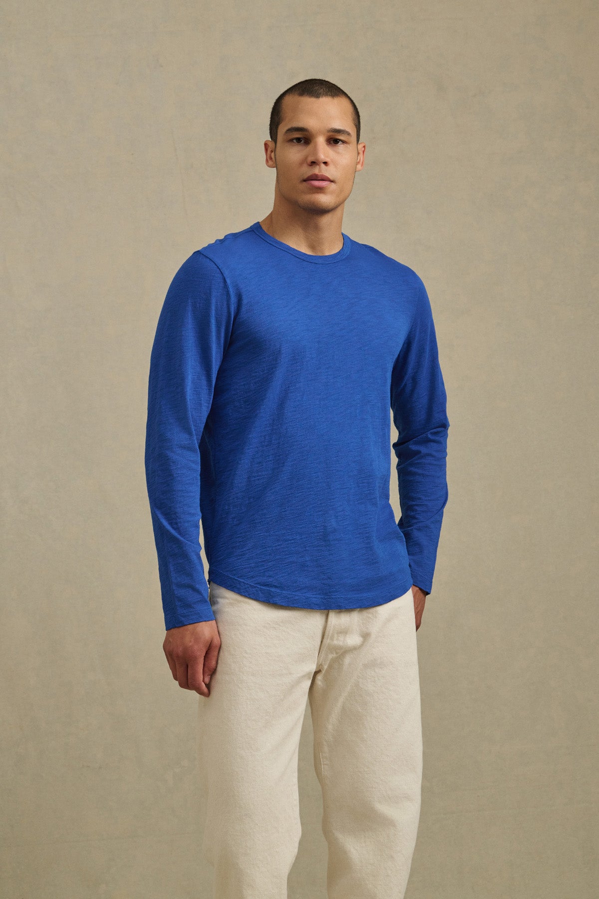 A man wearing the KAI TEE by Velvet by Graham & Spencer with a crew neckline and off-white pants stands against a plain, light-colored background.