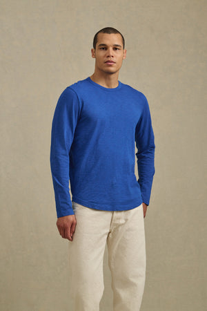 A man wearing the KAI TEE by Velvet by Graham & Spencer with a crew neckline and off-white pants stands against a plain, light-colored background.