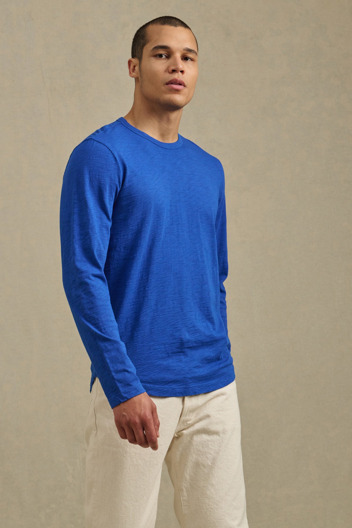 A person wears the KAI TEE by Velvet by Graham & Spencer, a bright blue Peruvian cotton long sleeve crewneck, paired with off-white pants against a plain beige background.