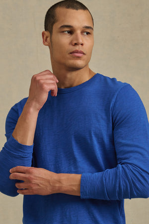 A man in a blue KAI TEE by Velvet by Graham & Spencer stands against a beige background, gazing to his left with one hand raised near his chin.
