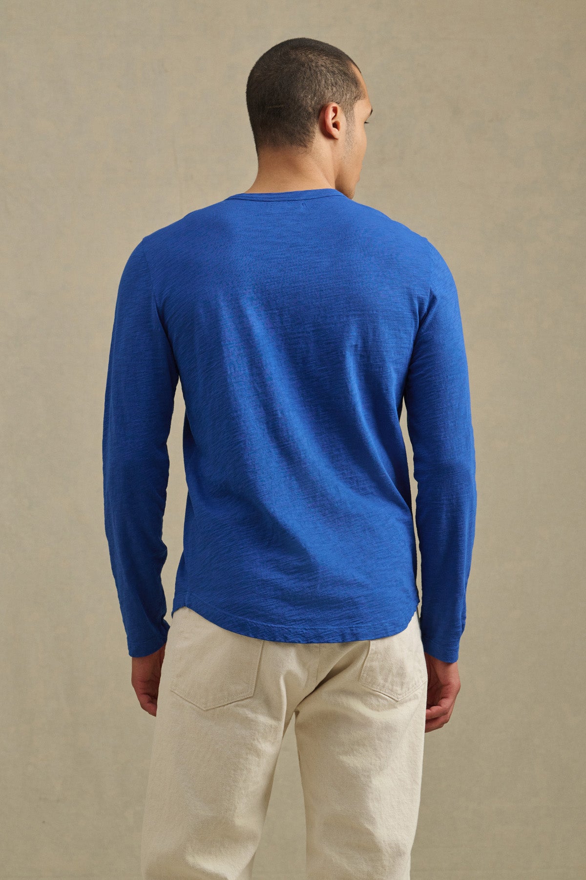 A person stands facing away, wearing the KAI TEE by Velvet by Graham & Spencer—a long-sleeve blue Peruvian cotton crewneck—paired with light pants against a neutral background.