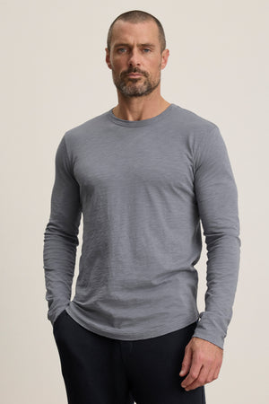 A man with short hair and a beard wears the light gray KAI TEE by Velvet by Graham & Spencer, featuring a Peruvian cotton long sleeve crew neck, paired with dark pants against a plain light background.