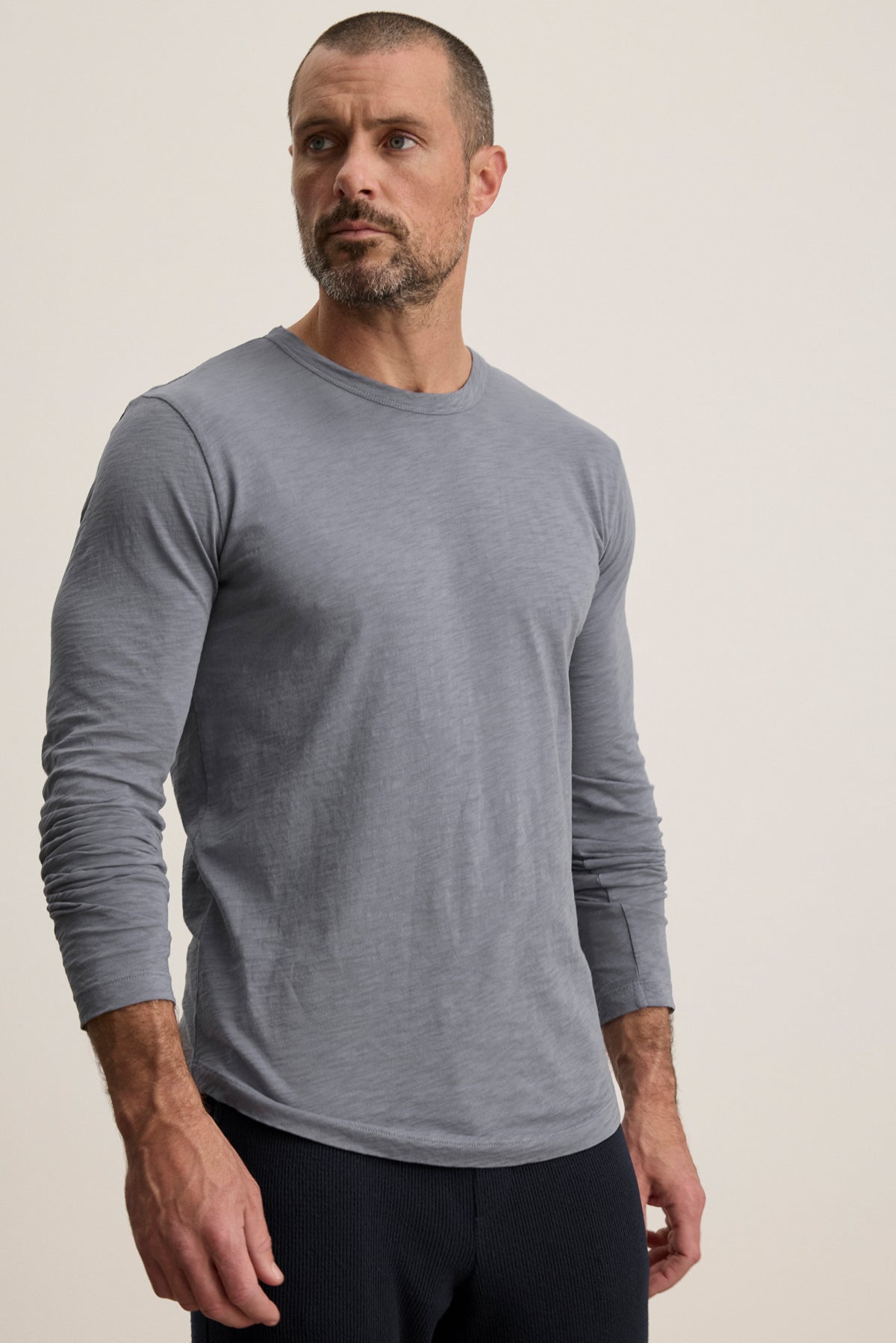 A man with short hair and a beard wears the KAI TEE by Velvet by Graham & Spencer—a light gray, long sleeve crewneck made from soft Peruvian cotton—paired with dark pants, standing against a simple light background.-40054343237825