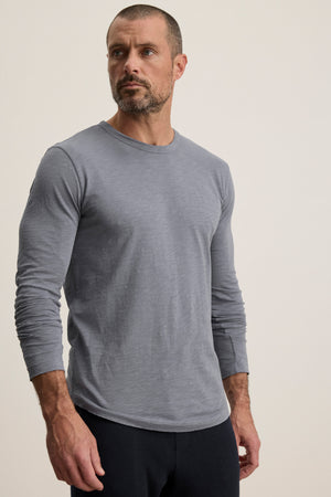 A man with short hair and a beard wears the KAI TEE by Velvet by Graham & Spencer—a light gray, long sleeve crewneck made from soft Peruvian cotton—paired with dark pants, standing against a simple light background.