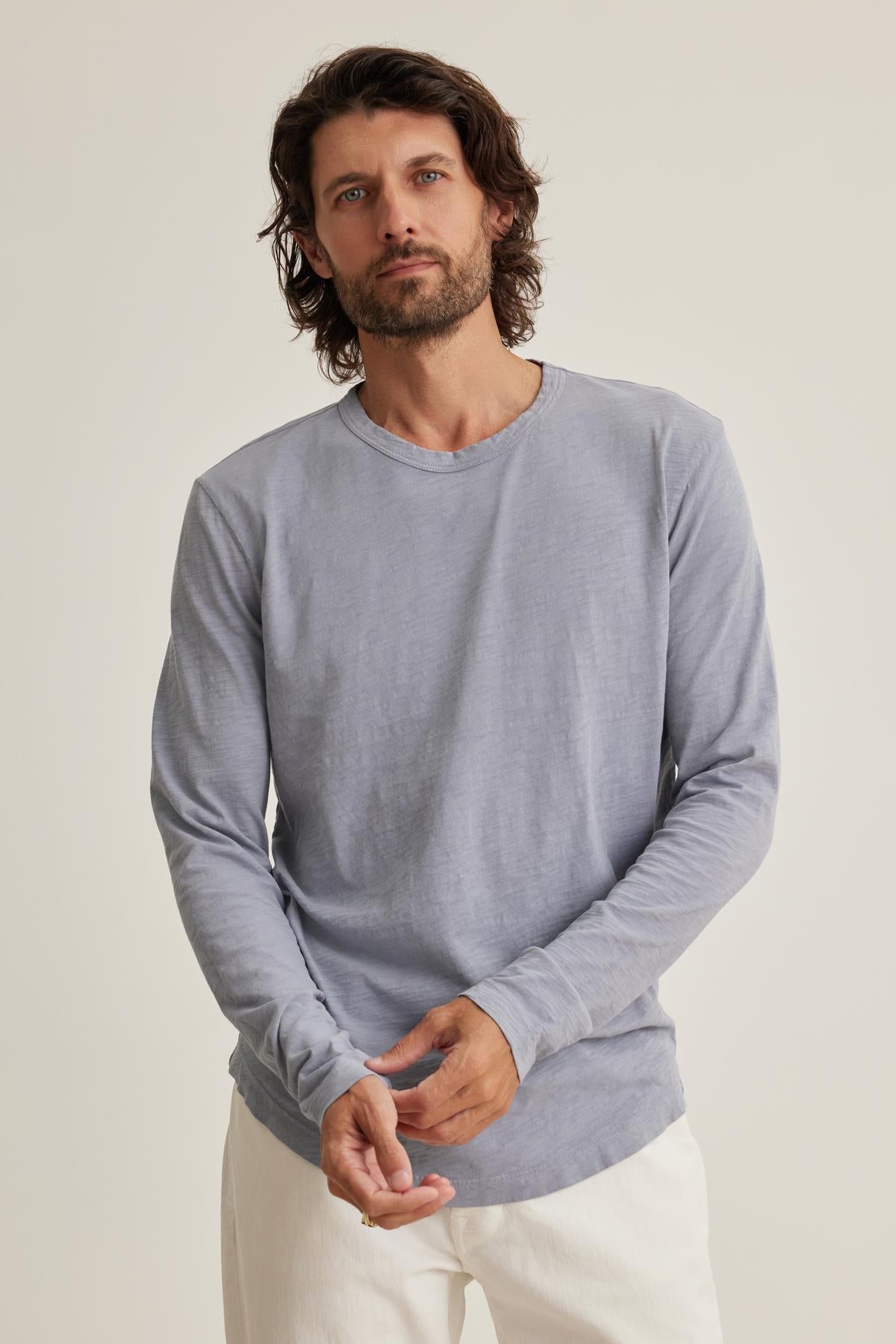   A man with shoulder-length hair and a beard wears the KAI TEE by Velvet by Graham & Spencer—a light gray long-sleeve shirt with a classic crew neckline—and white pants, standing against a plain background. 