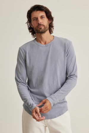 A man with shoulder-length hair and a beard wears the KAI TEE by Velvet by Graham & Spencer—a light gray long-sleeve shirt with a classic crew neckline—and white pants, standing against a plain background.