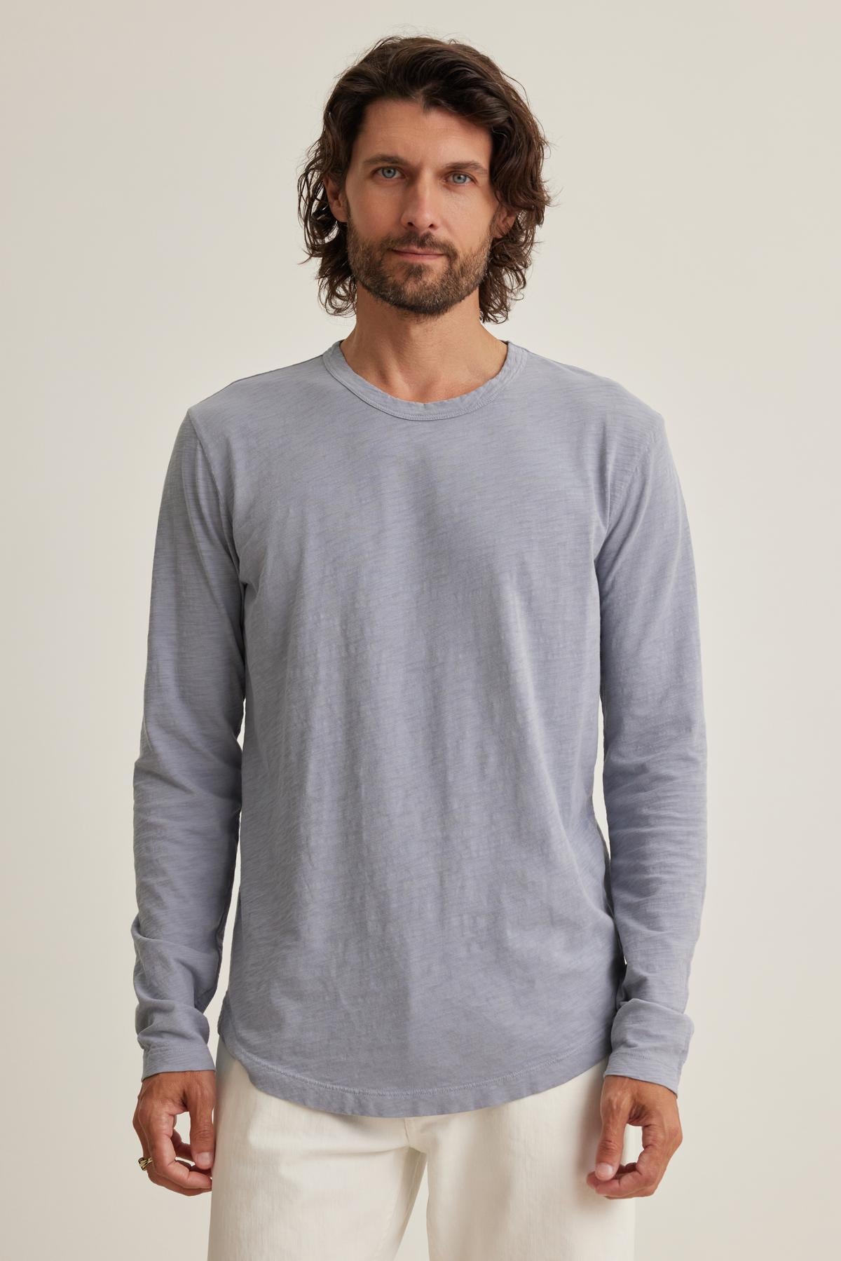   A man with wavy hair and a beard wears the KAI TEE by Velvet by Graham & Spencer, a light blue long-sleeve shirt with a classic crew neck, paired with white pants and standing against a plain background. 