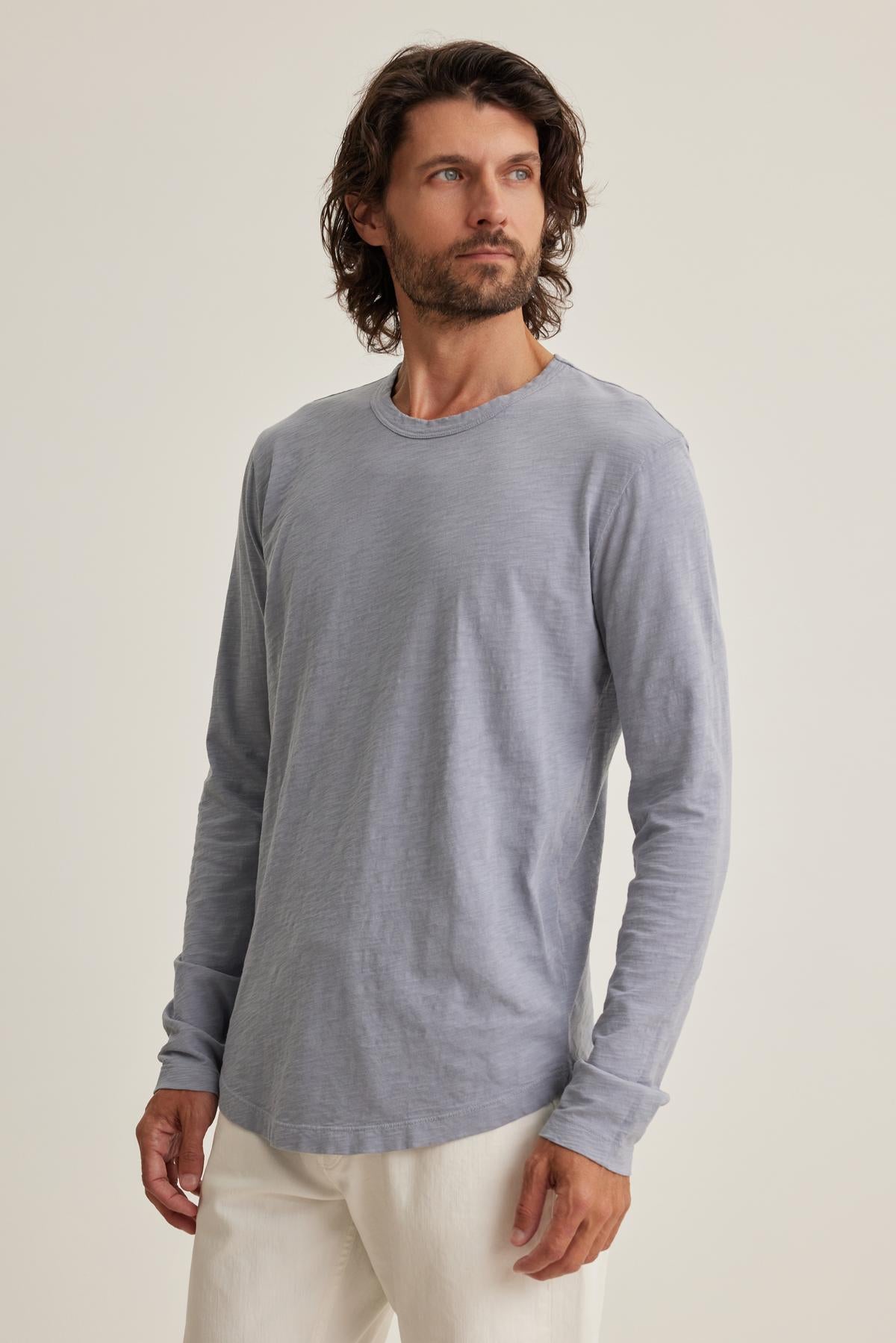 Wearing Velvet by Graham & Spencer's KAI TEE, featuring a gray long-sleeve and classic crew neckline, paired with white pants, a person stands against a plain background, gazing to the side.-38643311083713