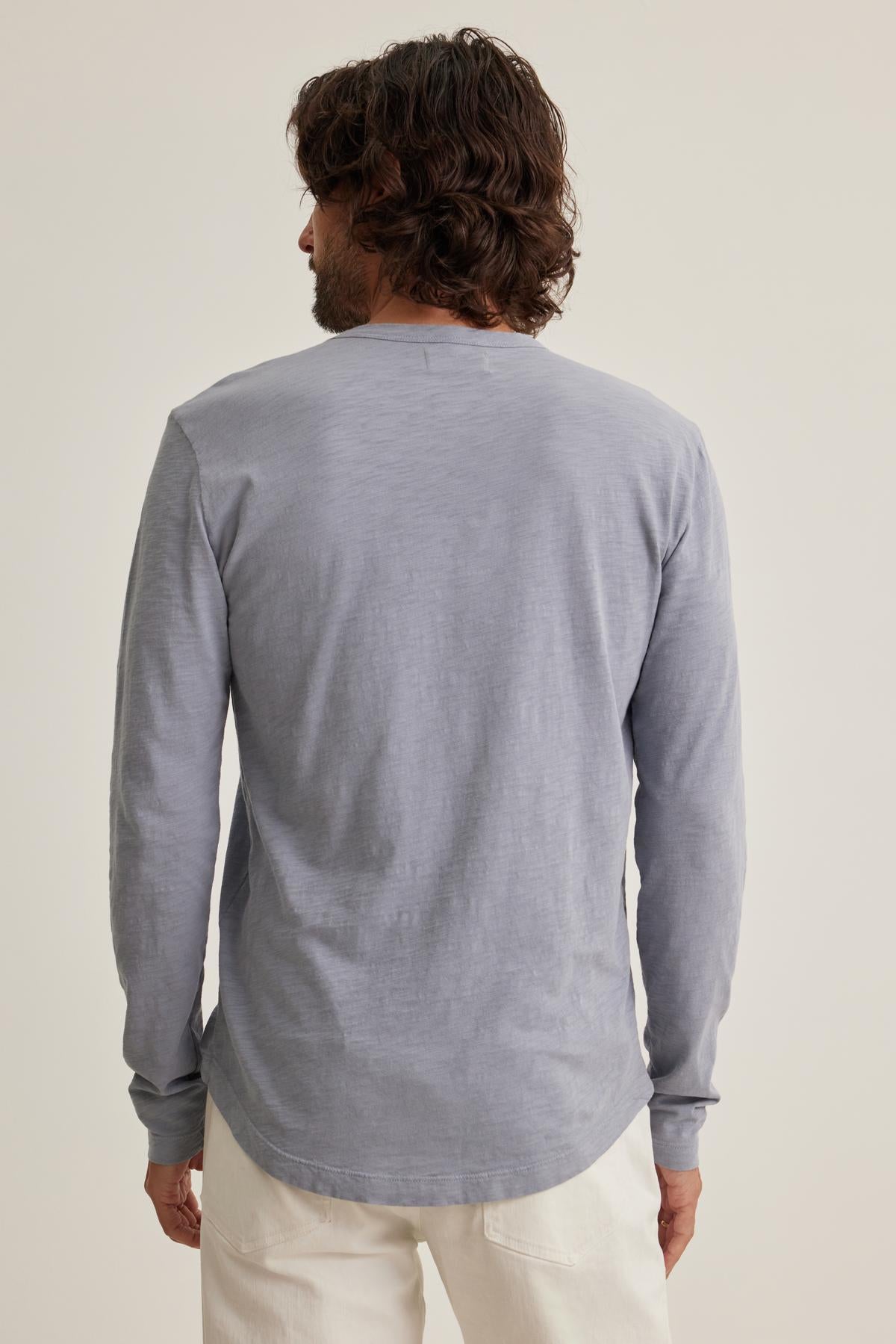 A person with long hair, seen from behind, dons the KAI TEE by Velvet by Graham & Spencer—a classic crew neck shirt in light gray—paired with white pants against a plain background, embodying timeless design.-38643308462273