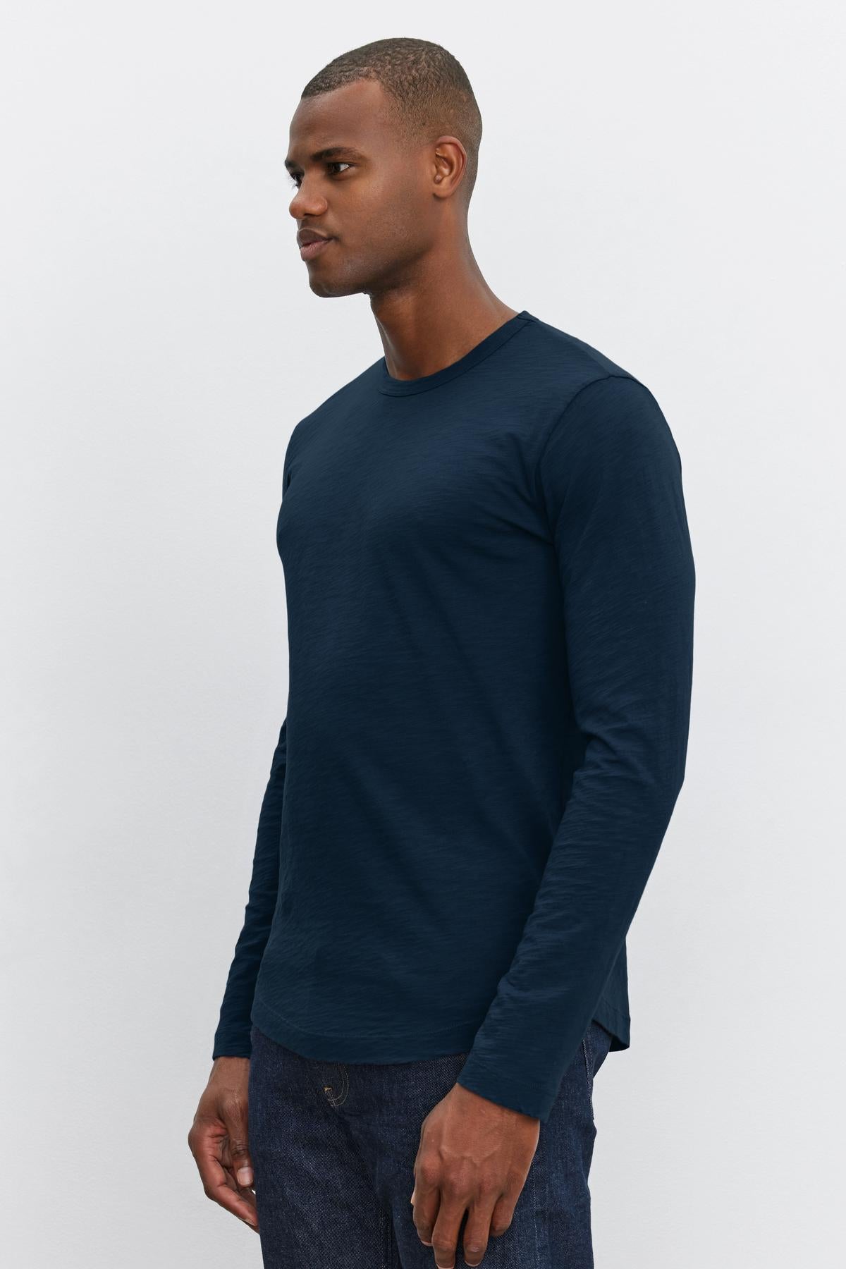 A man wearing a plain, long-sleeve, dark blue KAI TEE by Velvet by Graham & Spencer with a modern curved hemline and dark pants stands against a plain white background.-37469162340545