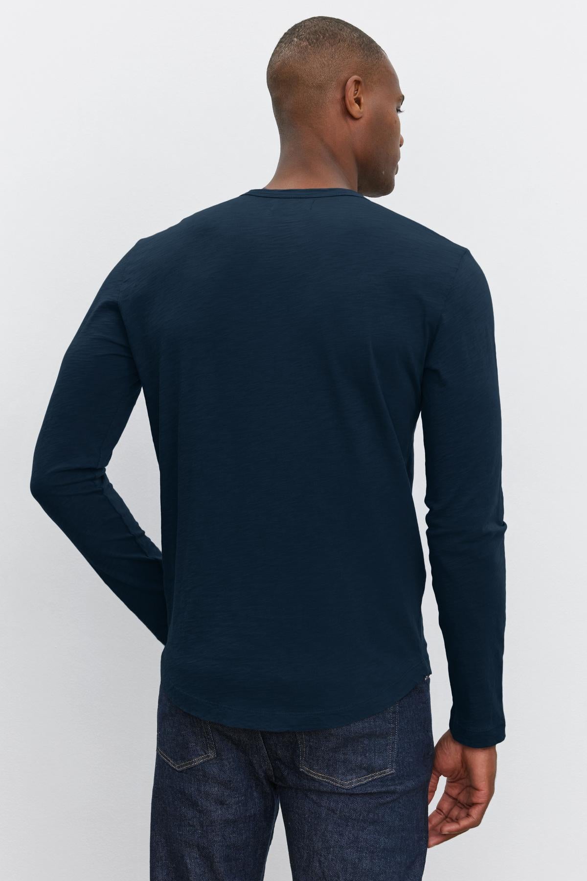   A person is shown from behind wearing a long-sleeve, dark blue KAI TEE by Velvet by Graham & Spencer with a classic crew neckline and dark denim jeans. 