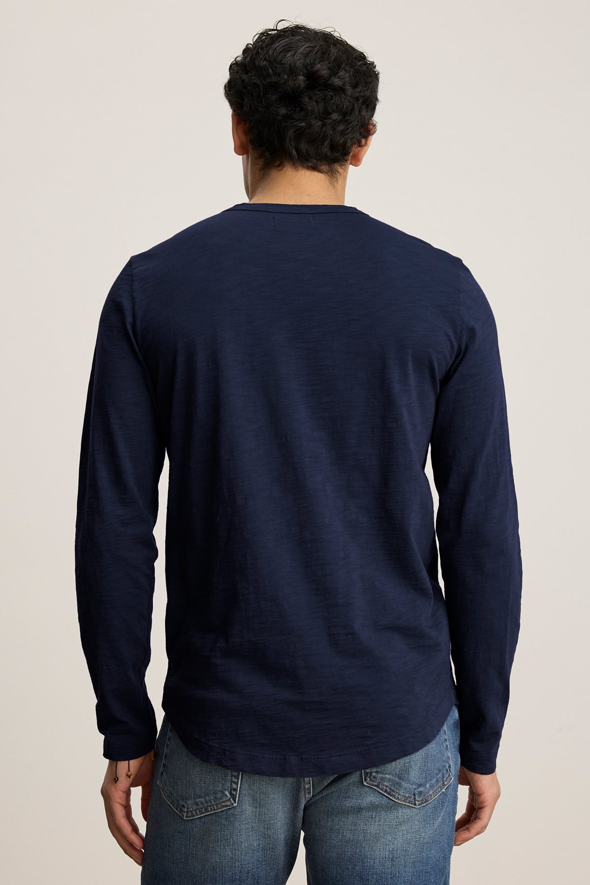   A man with curly hair, wearing the KAI TEE by Velvet by Graham & Spencer in navy, crafted from luxurious Peruvian cotton with a classic crew neckline, paired with jeans, faces away from the camera. 