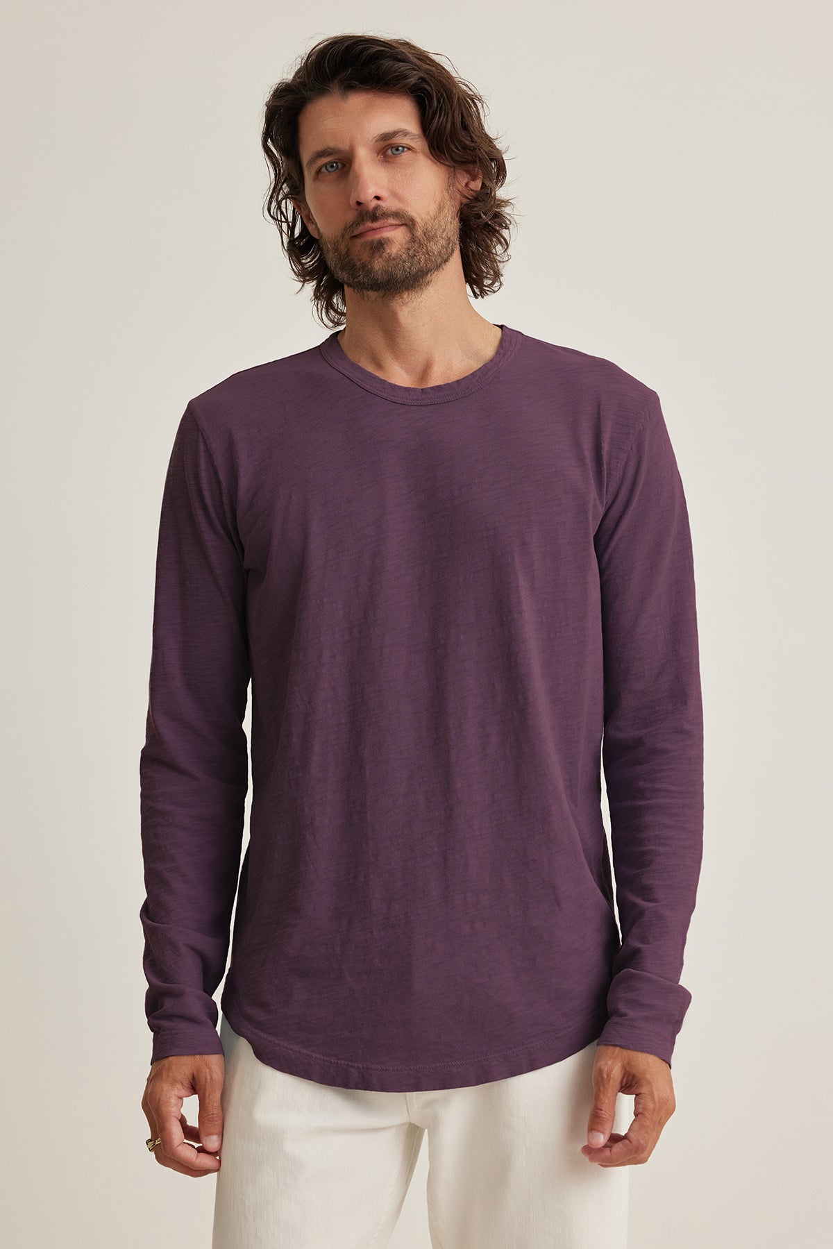   A man with long hair wears the KAI TEE by Velvet by Graham & Spencer, a purple long-sleeve shirt made of soft Peruvian cotton, paired with crisp white pants. Against a plain background, he exudes effortless elegance highlighted by the shirt's classic crew neckline. 