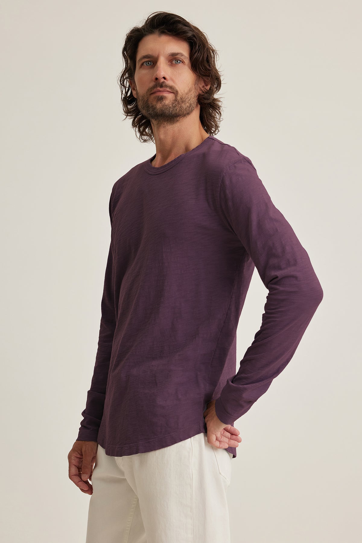 A man with long hair wears Velvet by Graham & Spencer's KAI TEE, a purple long-sleeve shirt paired with white pants, exuding timeless design as he gazes slightly upward against a plain background.-38807449239745