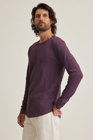 A man with long hair wears Velvet by Graham & Spencer's KAI TEE, a purple long-sleeve shirt paired with white pants, exuding timeless design as he gazes slightly upward against a plain background.