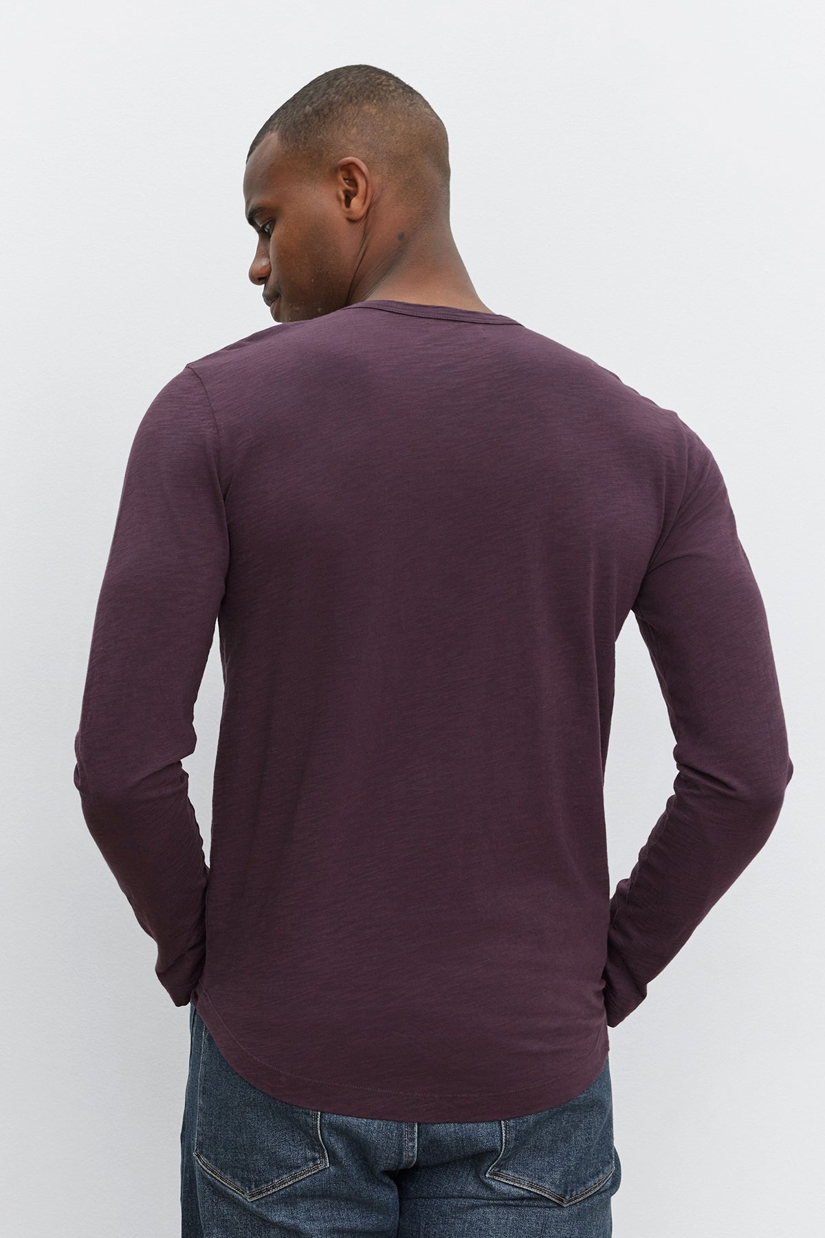   A man stands facing away from the camera, wearing a KAI TEE long-sleeved dark purple shirt made from Peruvian cotton by Velvet by Graham & Spencer and jeans against a plain white background. 