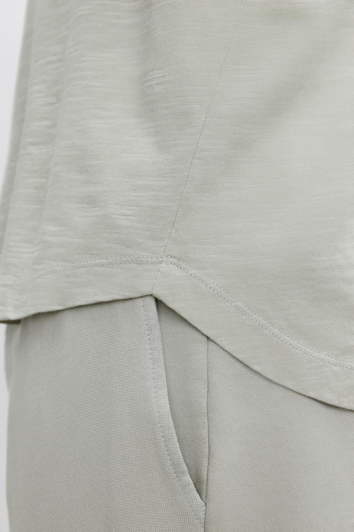   Close-up of a light grey fabric shirt over matching light grey pants. The KAI TEE by Velvet by Graham & Spencer, made from textured cotton slub, features visible seam details and a modern curved hemline. 