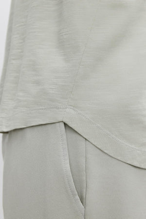 Close-up of a light grey fabric shirt over matching light grey pants. The KAI TEE by Velvet by Graham & Spencer, made from textured cotton slub, features visible seam details and a modern curved hemline.