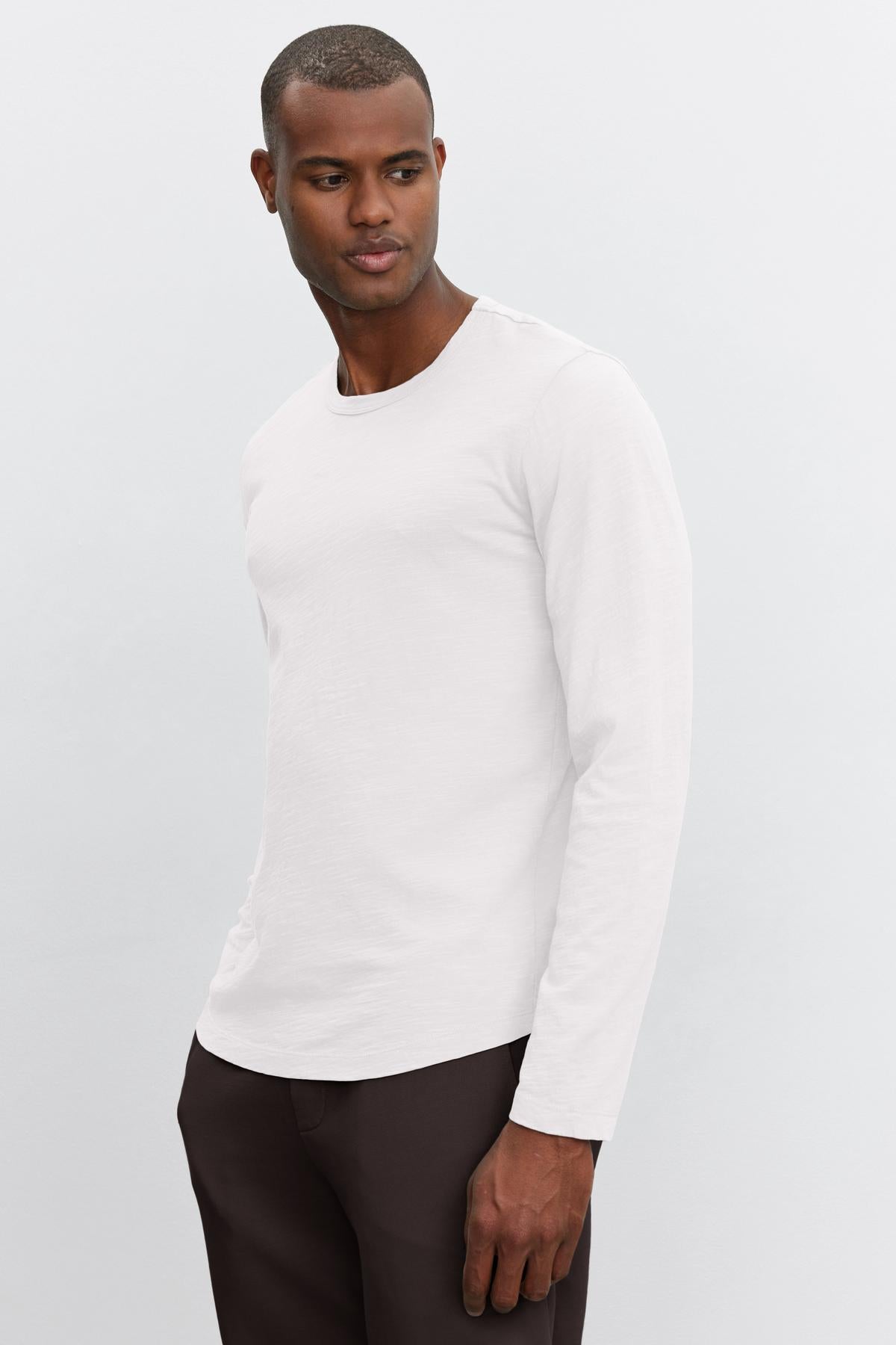A man in a white long-sleeve slub knit shirt and dark pants standing against a light grey background, looking to his right, wearing the Velvet by Graham & Spencer KAI CREW NECK TEE.-36544896958657