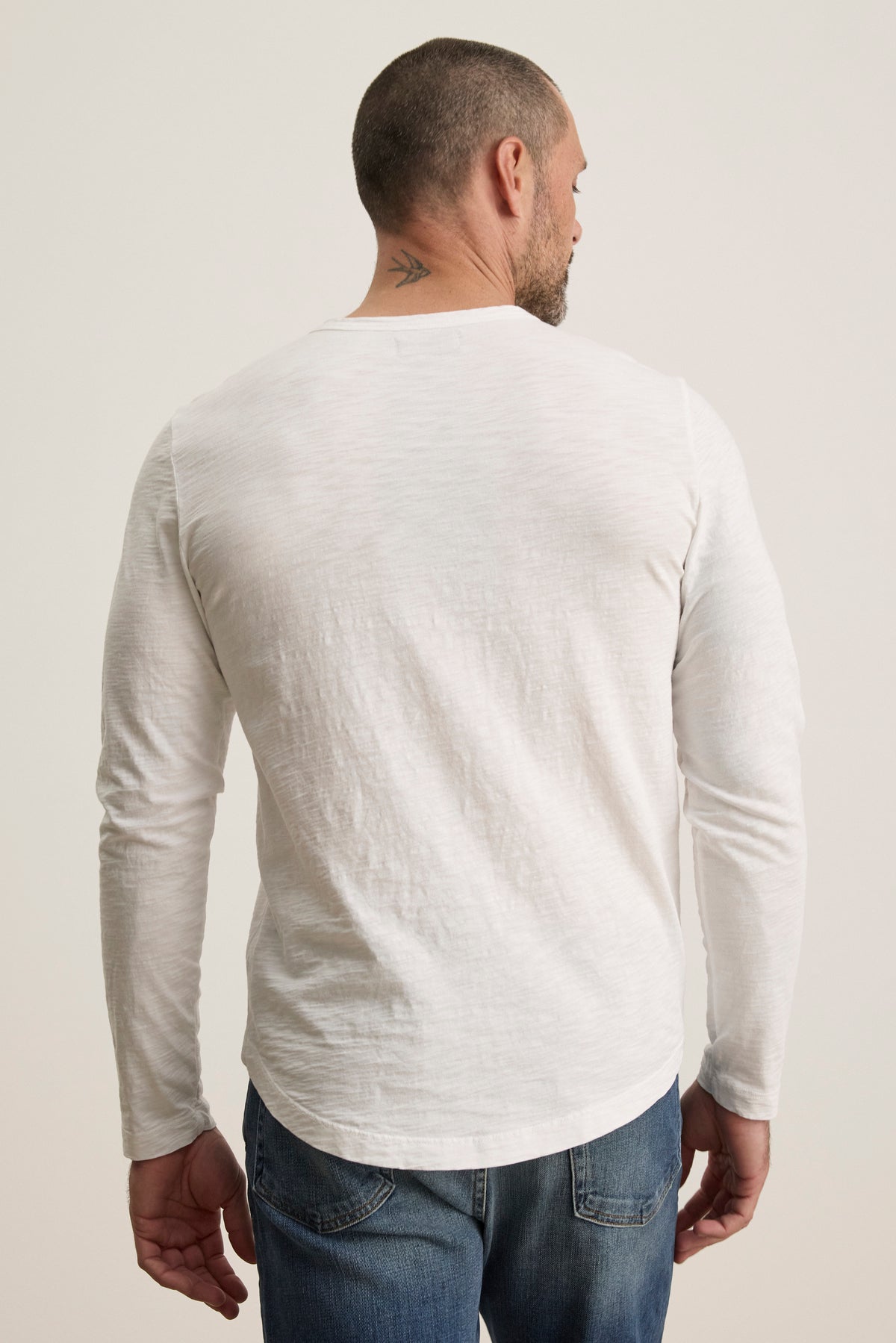   A man wearing Velvet by Graham & Spencer's KAI TEE, a white long-sleeve shirt made from luxurious Peruvian cotton with a classic crew neckline, turns away, showcasing a small neck tattoo. 