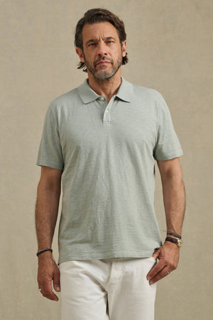 Man with short brown hair and beard wears the Velvet by Graham & Spencer MORRIS POLO in light green, featuring short sleeves and a ribbed collar, paired with white pants, standing against a plain beige background.