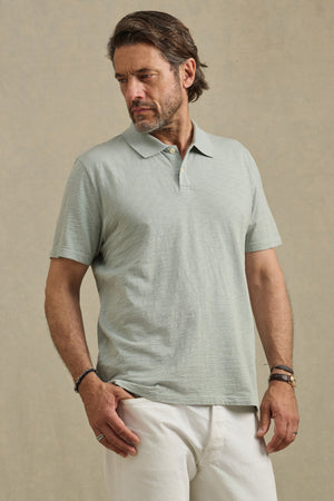 A man wearing the MORRIS POLO by Velvet by Graham & Spencer—a light gray cotton slub polo with a ribbed collar and short sleeves—pairs it with white pants, standing against a neutral background and looking left with one hand in his pocket.